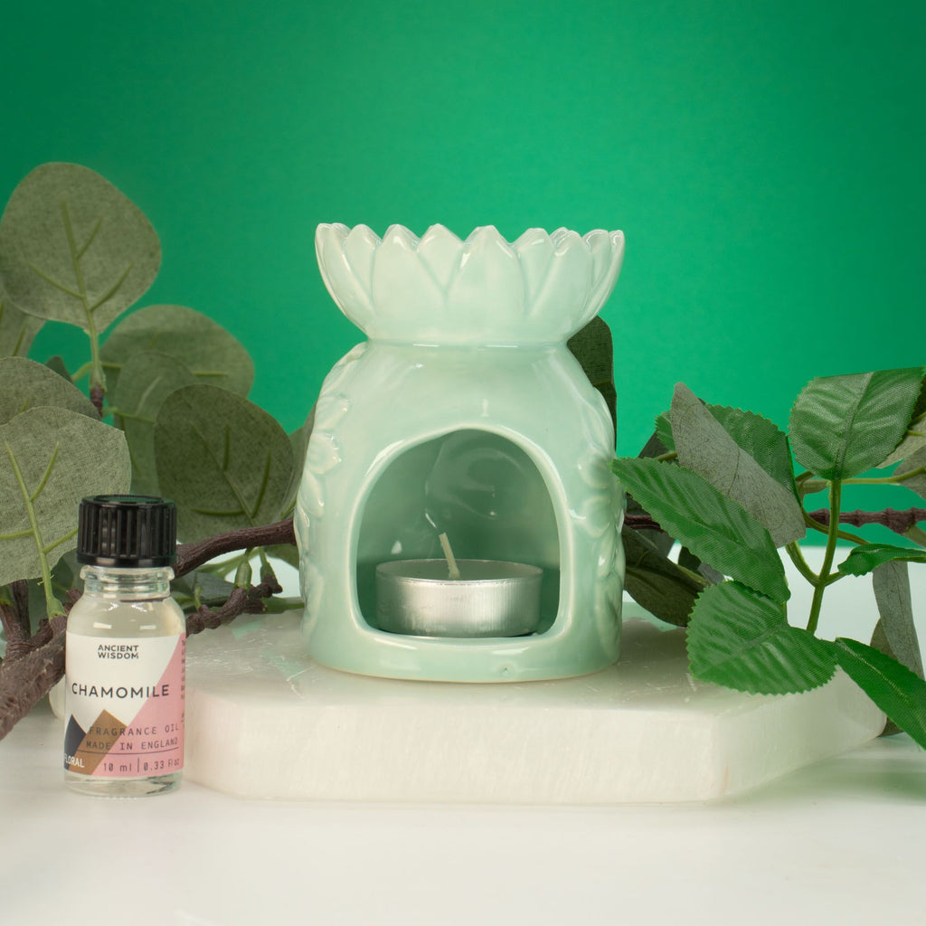 Mint green wax warmer with a small candle inside, next to a bottle of chamomile essential oil on a white surface with green leaves in the background.