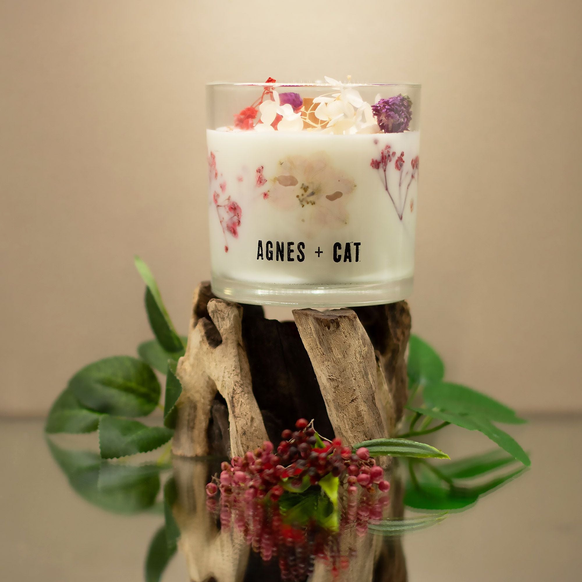 Candle in a glass holder with floral design on a wooden stand, featuring the brand 'AGNES + CAT'.