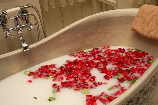 Bath tub filled with bubbles and red rose petals, with a towel and faucet in the background.