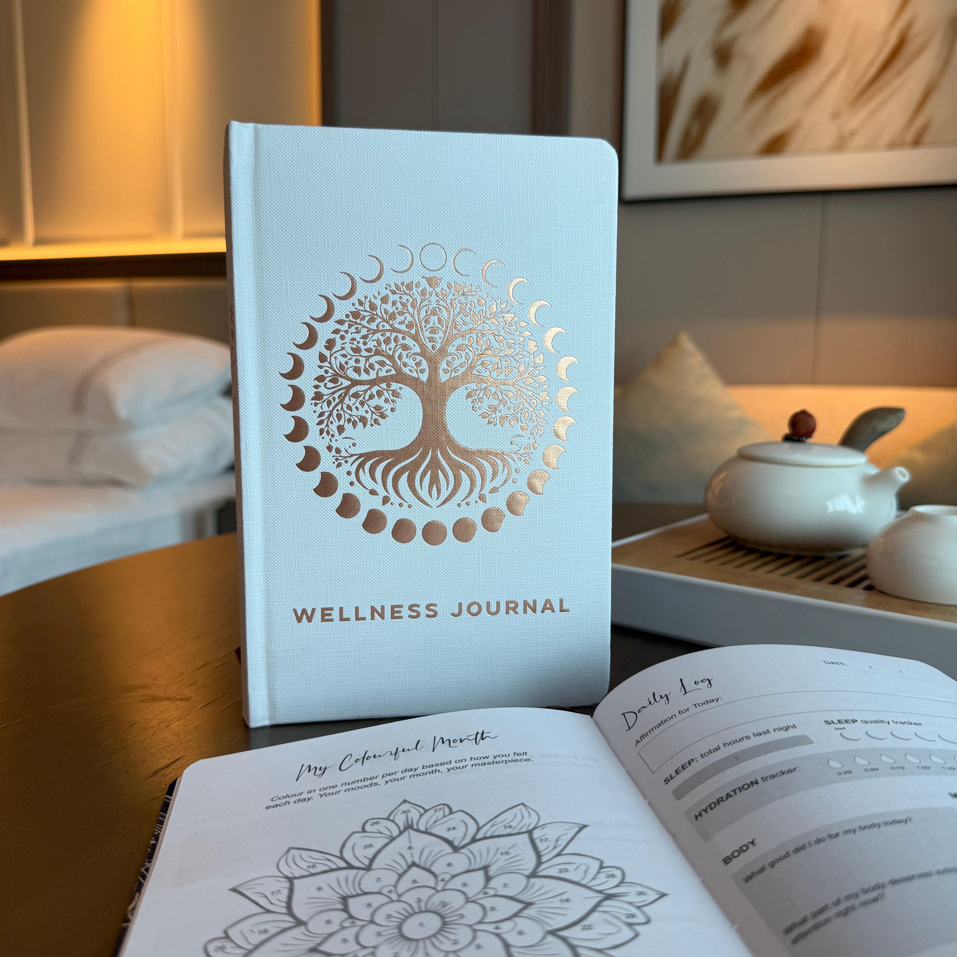Wellness journal with tree of life design on cover, placed on a table with a tea set in the background.